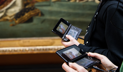 Two people hold Nintendo devices connected to headphones as they learn about an artwork from the Lourve.