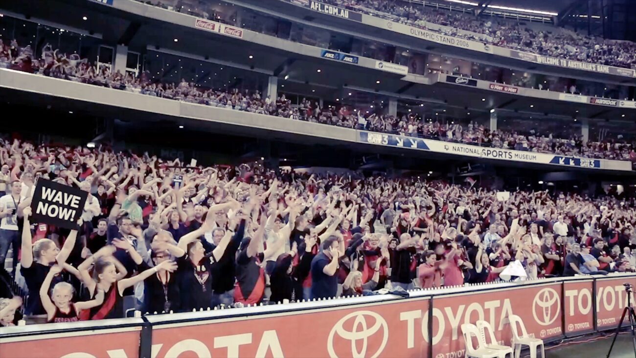 Crowd of AFL fans holds ‘Wave Now’ signs while waving their arms as part of Visa Wave and Win.