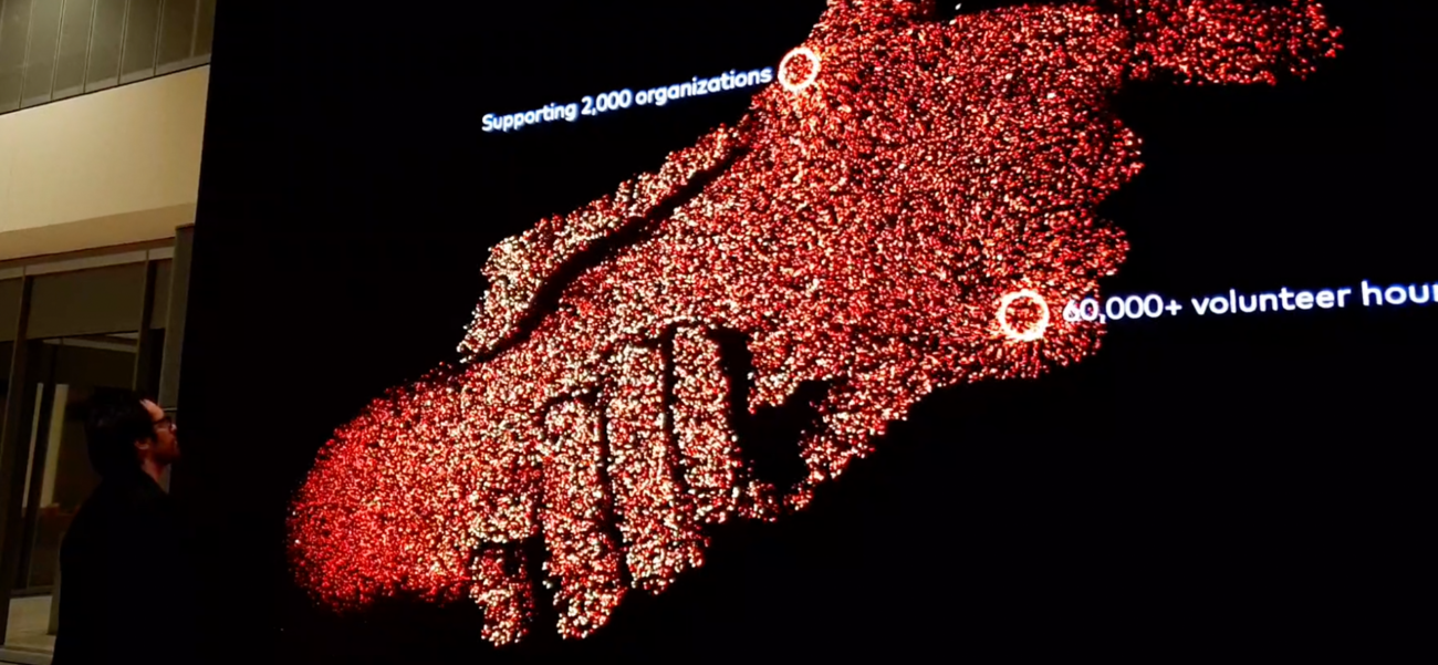 Iain Greenhalgh from S1T2 looks up at particle data visualisation showing two hands grasping one another on a large-scale LED screen.