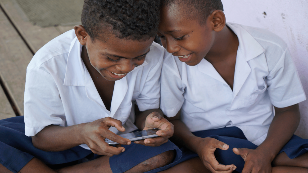 Two Fijian children play Beyond the Stars mobile game on a smartphone during school.